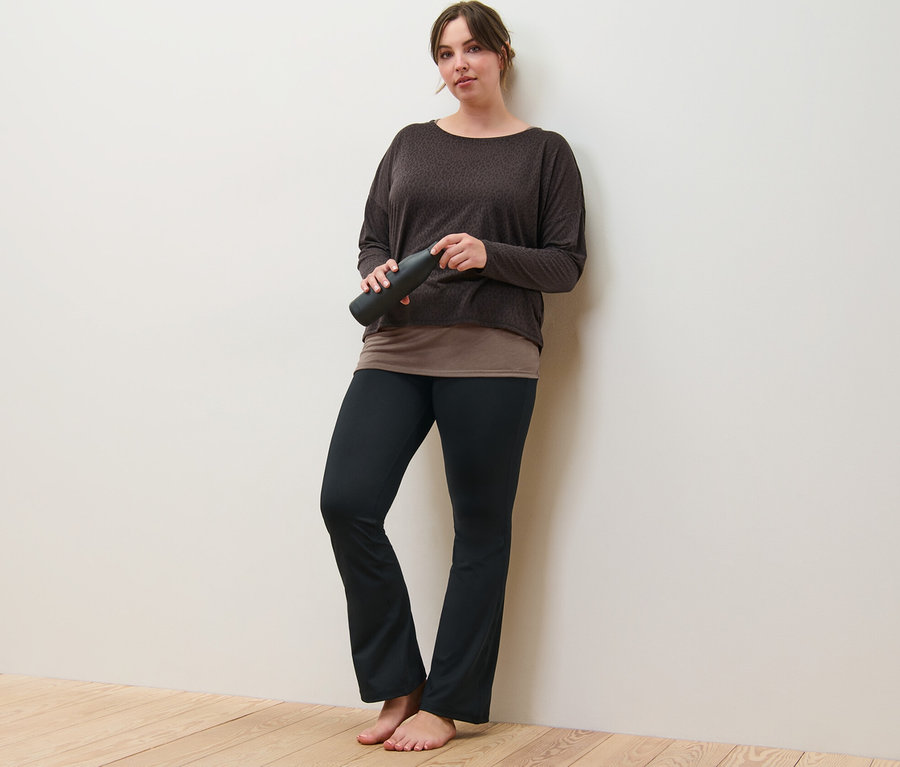 Une femme appuyée contre un mur tenant un t-shirt de sport 2 en 1 marron et un pantalon de sport modelant.