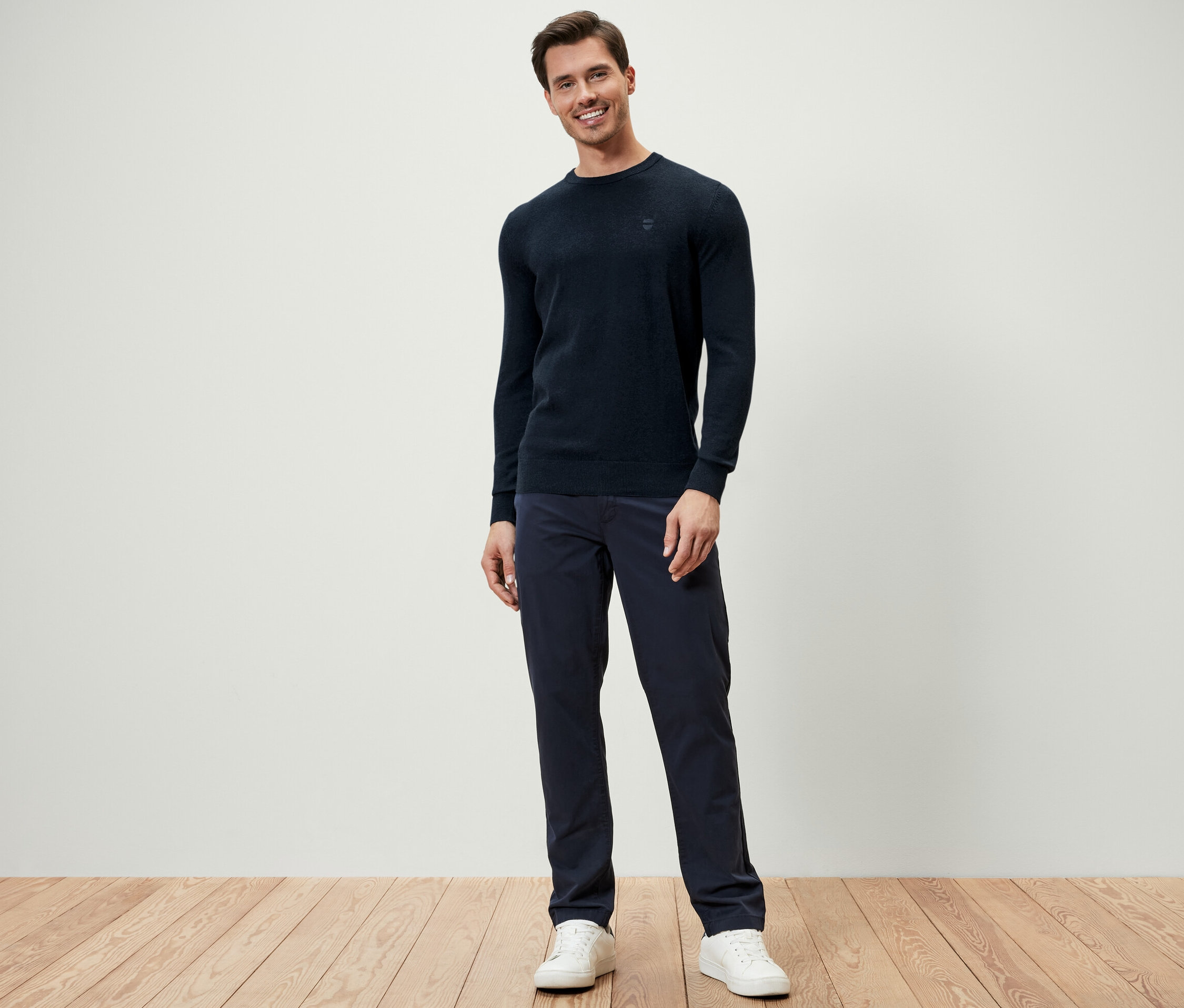 Un homme debout portant un pull à encolure ronde bleu foncé, un pantalon bleu foncé et des baskets blanches.