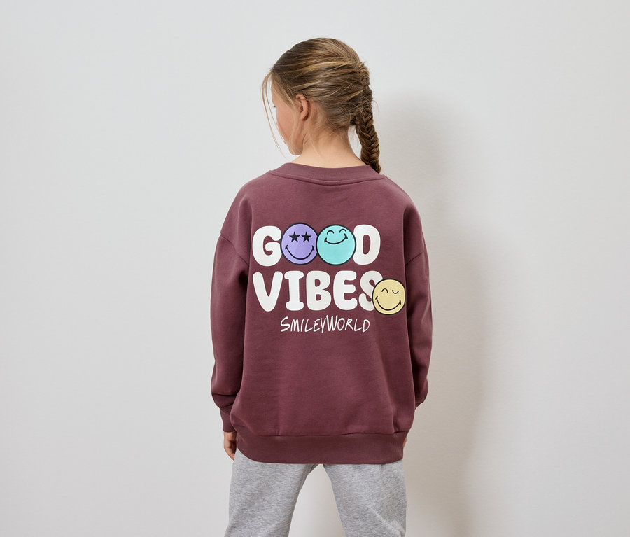 Une fille, vue de dos, porte un sweat-shirt bordeaux avec l'inscription Good Vibes Smileyworld.