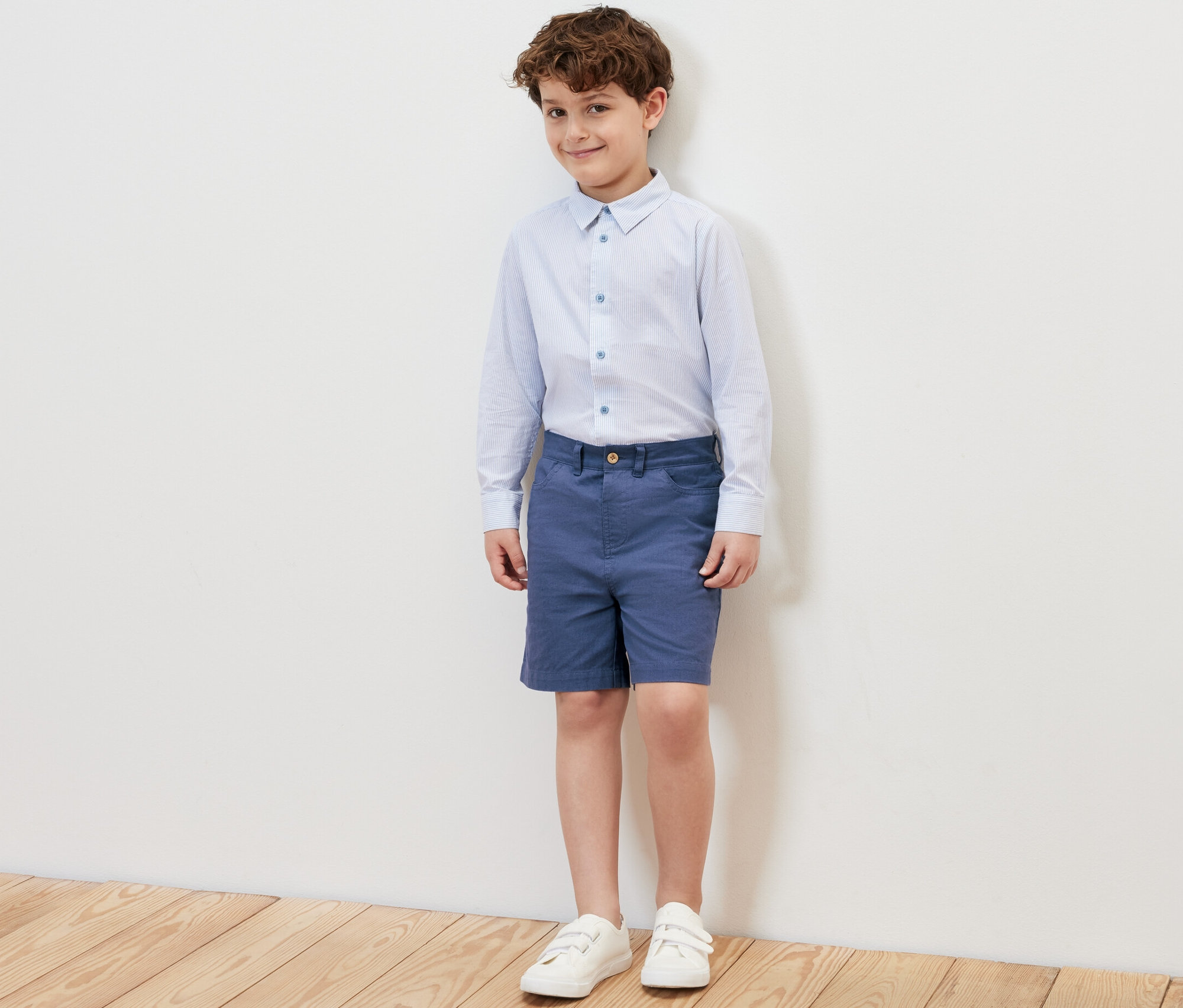 Un garçon debout portant un short pour enfant bleu, une chemise et des baskets.