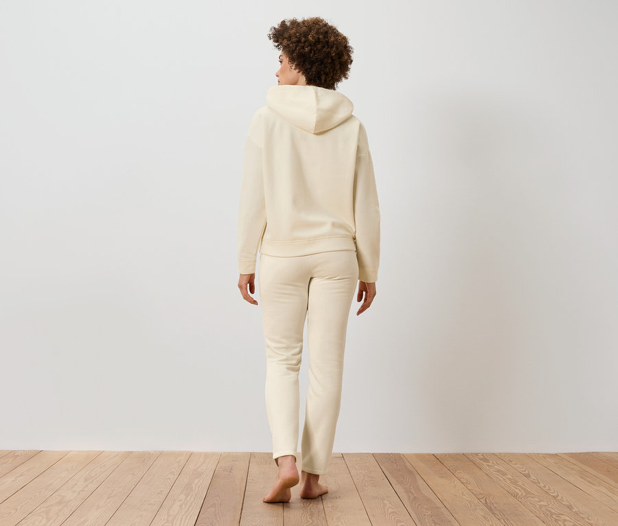Une femme aux cheveux bouclés, vue de dos, porte un sweat à capuche beige et un pantalon de jogging sur un plancher en bois.