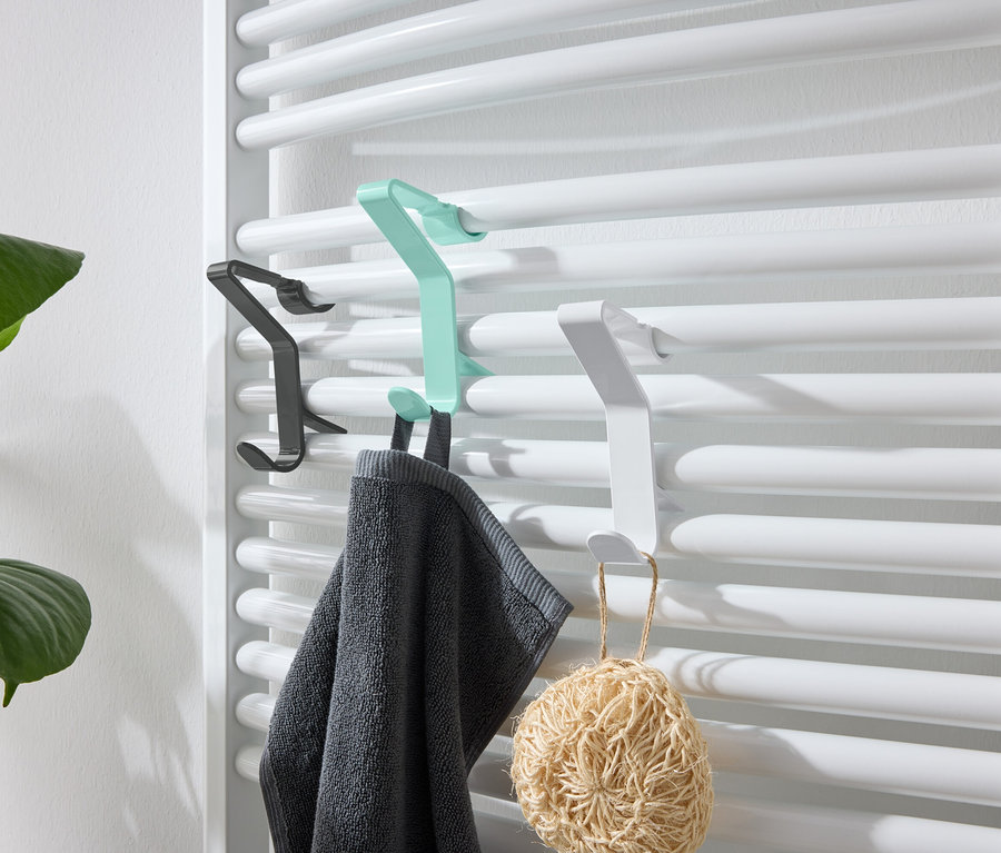 Des crochets noirs, verts et blancs sont accrochés à un radiateur blanc. Une serviette grise est accrochée au crochet vert et un luffa au crochet blanc.