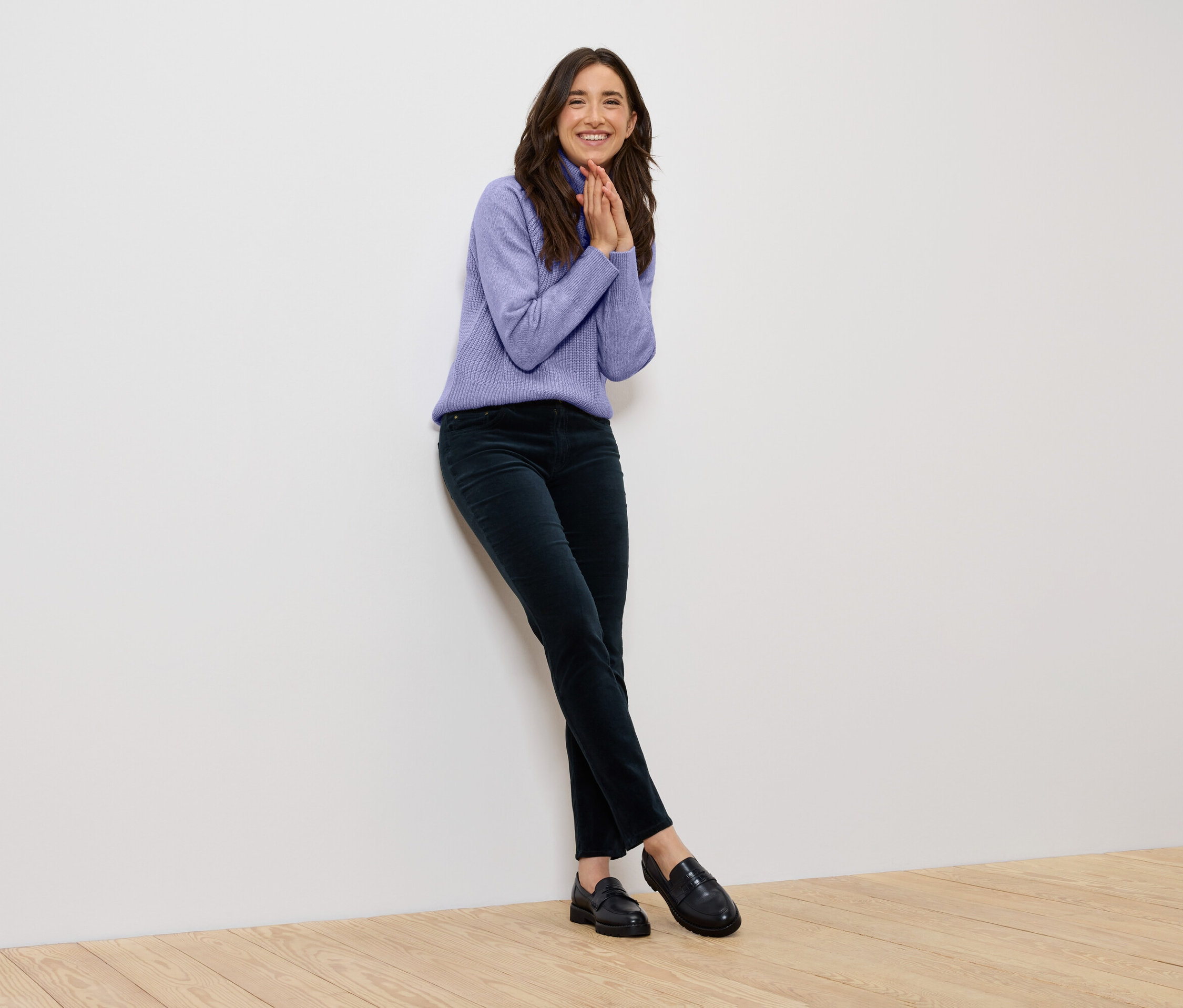 Une femme souriante portant un pull violet et un pantalon vert foncé, appuyée contre un mur blanc.