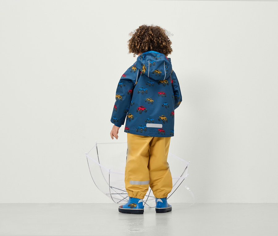 Un garçon, dos à la caméra, porte une veste imprimée avec des voitures et un pantalon jaune à côté d'un parapluie transparent.