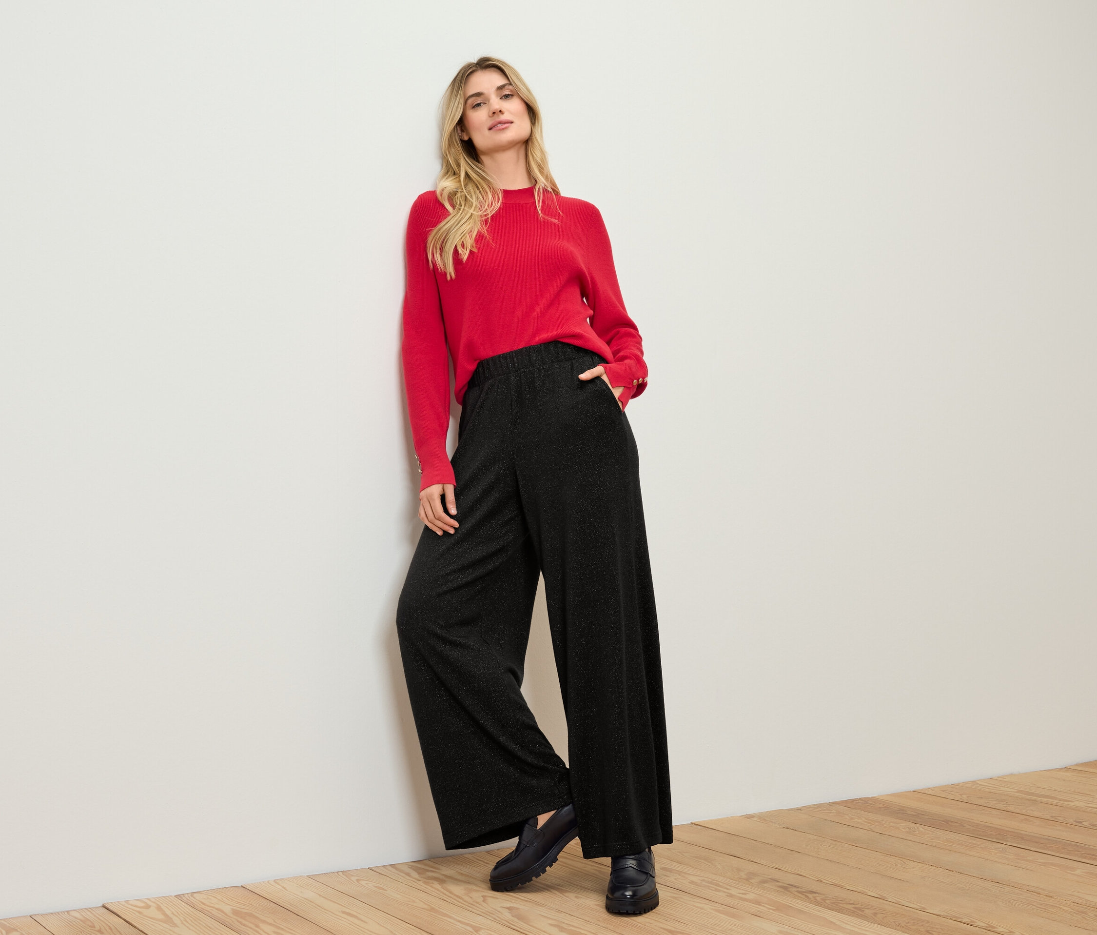 Blonde en pull rouge et pantalon noir appuyée contre un mur blanc.