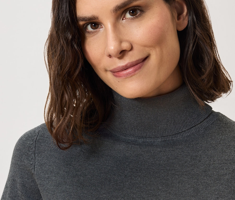 Gros plan sur une femme souriante aux cheveux bruns portant un pull à col roulé gris.
