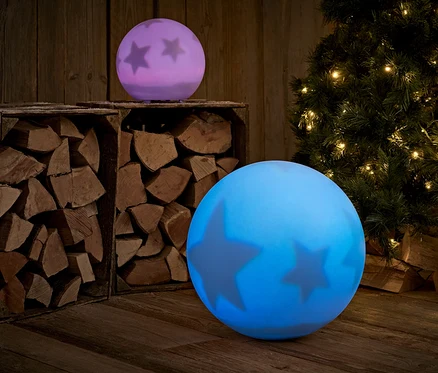 Deux boules lumineuses avec des étoiles se trouvent sur un plancher en bois, avec du bois et un arbre de Noël en arrière-plan.