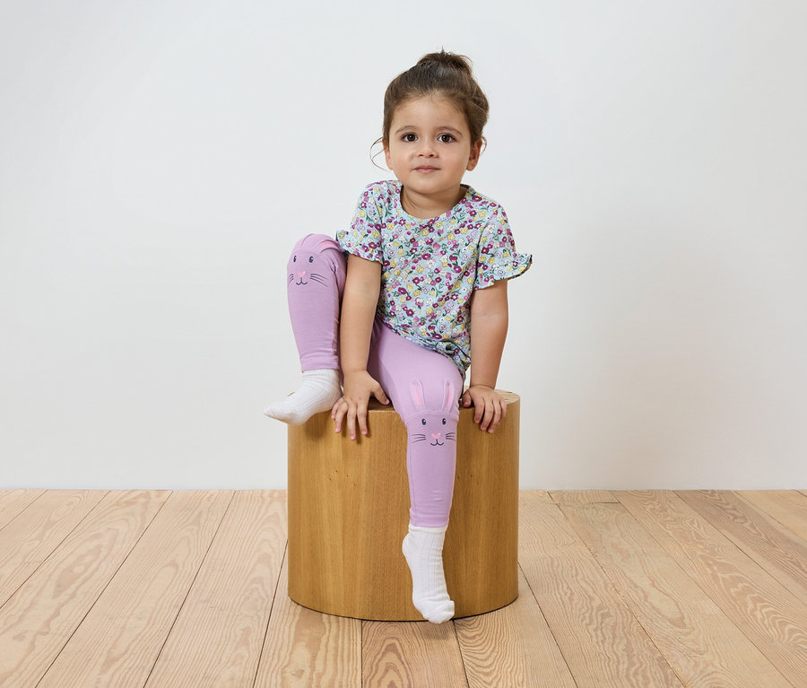 Une fillette est assise sur un tabouret en bois. Elle porte un t-shirt à fleurs et un legging violet avec un motif de chat.
