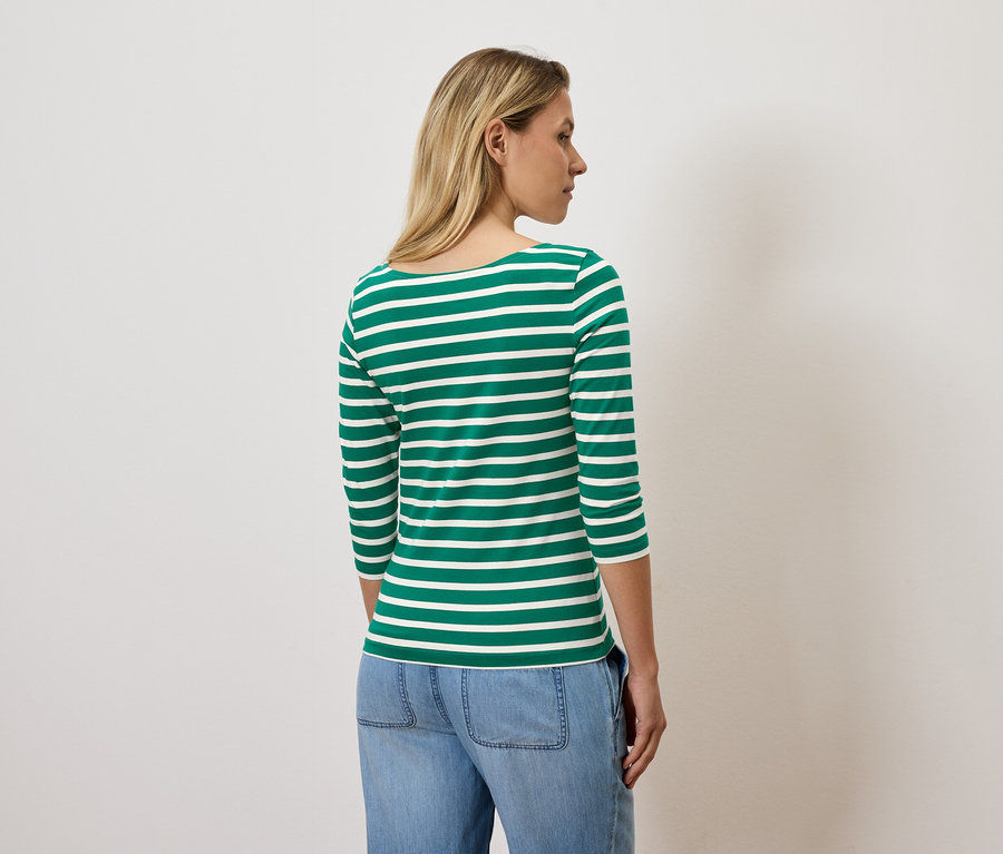 Une femme aux cheveux blonds se tient dos à la caméra et porte un t-shirt à manches trois-quarts rayé vert et blanc et un jean bleu.