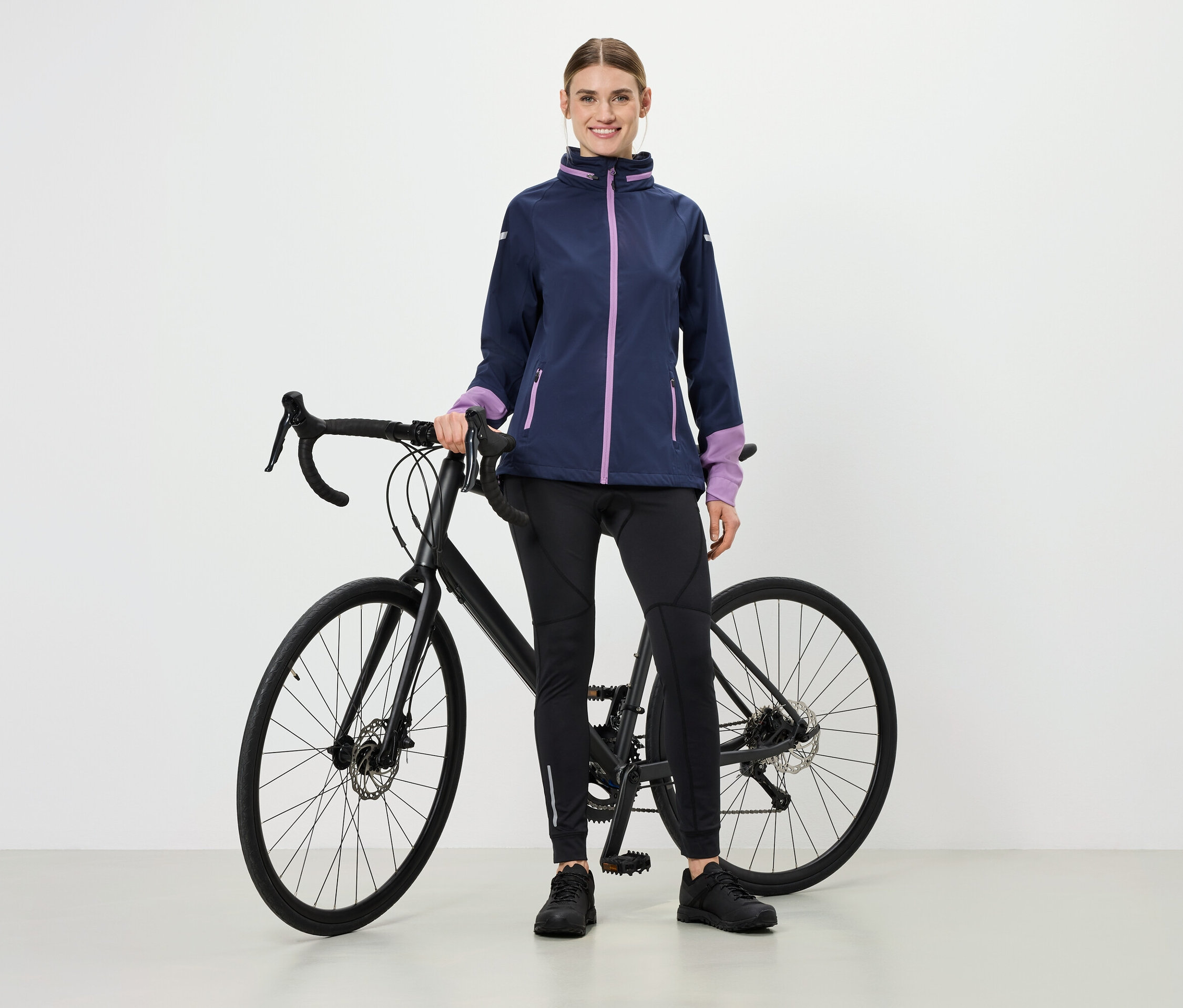 Une femme se tient à côté d'un vélo, elle porte une veste avec des accents violets, un legging de sport noir et des chaussures.