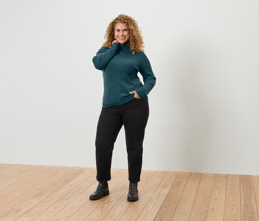 Une femme aux cheveux bouclés pose en pull col roulé vert, pantalon noir et bottes.