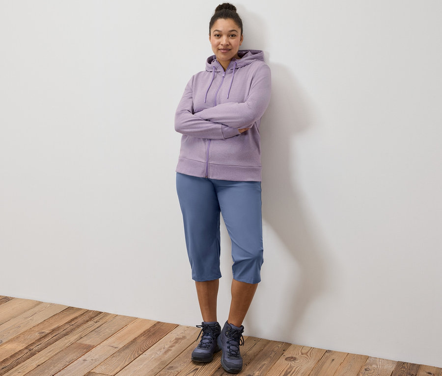 Une femme se tient debout, les bras croisés, portant un sweat à capuche violet et un Capri fonctionnel.