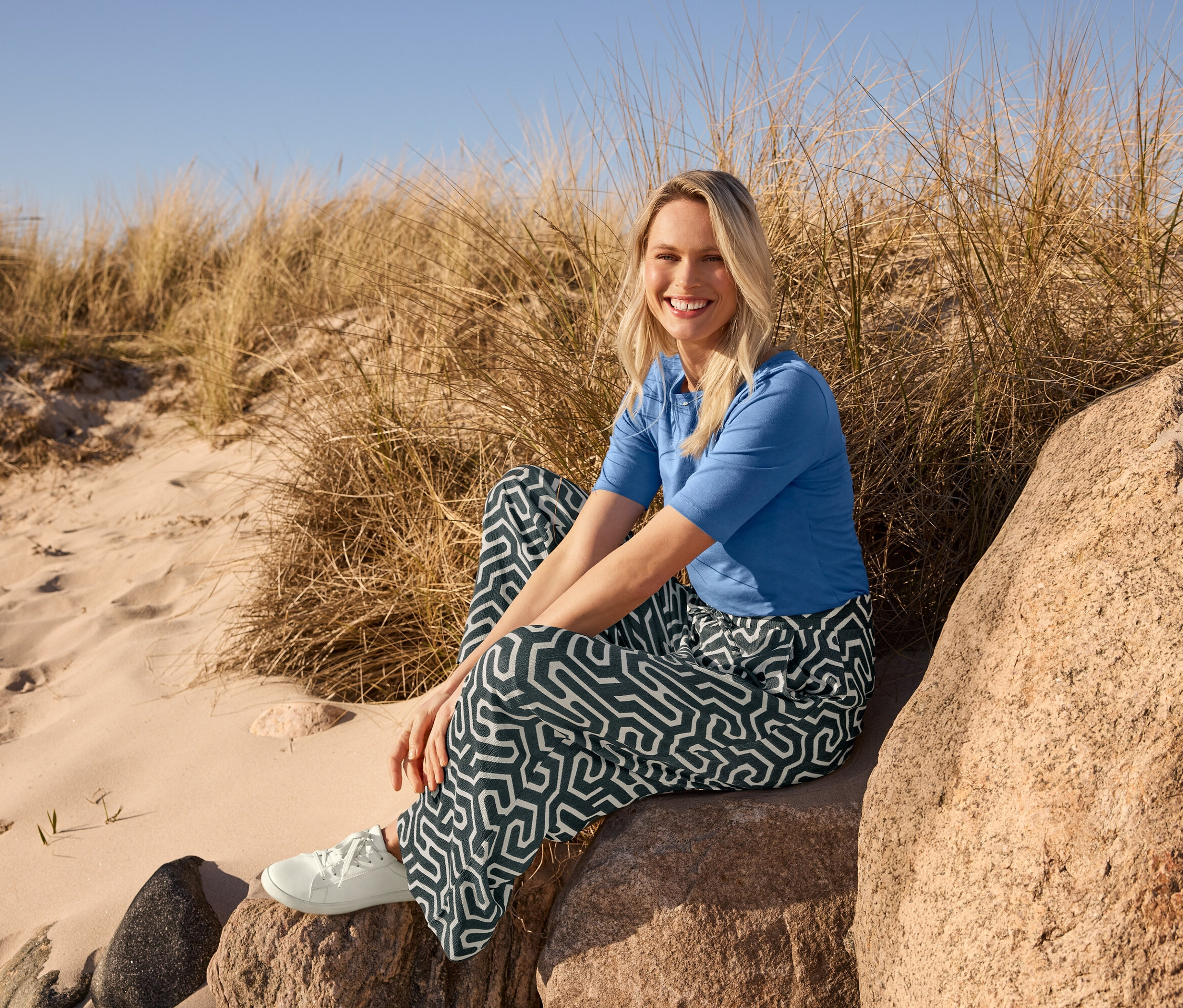 Une femme assise sur une pierre à la plage porte un t-shirt demi-manches bleu, un pantalon imprimé à enfiler avec effet Crinkle et des sneakers en cuir.