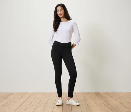 Une femme se tient debout, les mains dans les poches d'un jean noir, un chemisier blanc et des baskets blanches.