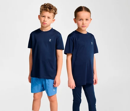 Deux enfants debout l'un à côté de l'autre. Le garçon à gauche porte un t-shirt d’entraînement HUMMEL HMLJR pulse bleu foncé et un short bleu. La fille à droite porte un t-shirt bleu foncé et un legging.
