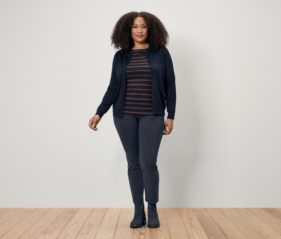 Une femme aux cheveux bouclés foncés pose sur un plancher en bois, portant un cardigan bleu foncé, un t-shirt rayé et un pantalon et des bottes bleu foncé.