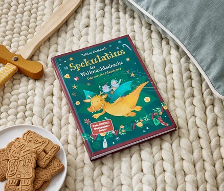 Un livre Spekulatius der Weihnachtdsrache repose sur une couverture tricotée à côté d'une assiette de pain d'épice et d'une épée en bois.