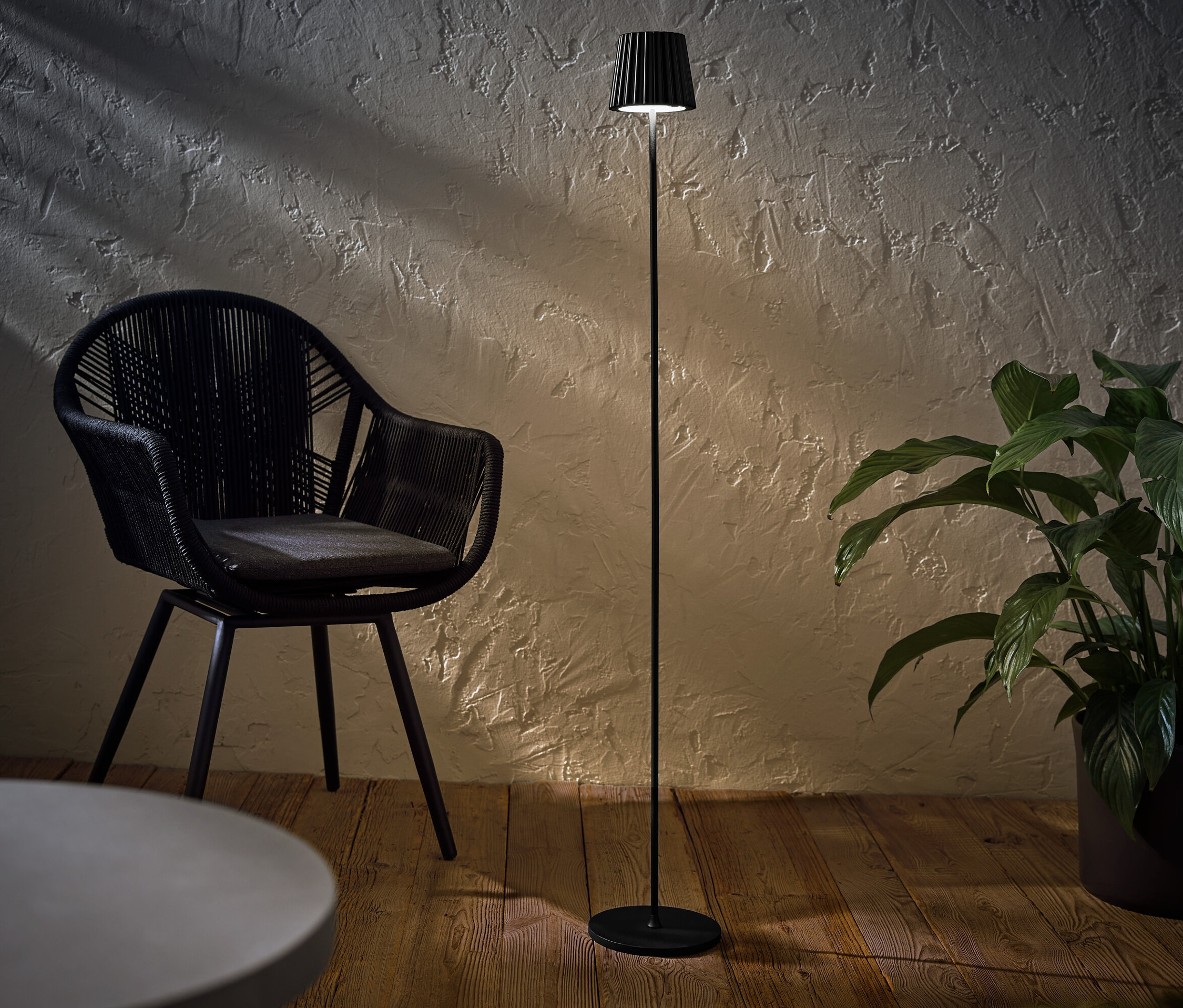 Un lampadaire d’extérieur à LED rechargeable, noir, se trouve à côté d’un fauteuil en osier noir et d’une plante en pot.