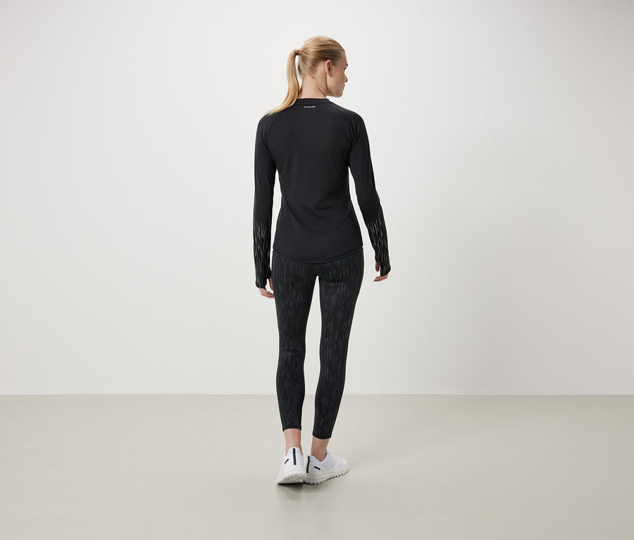 Une femme, dos à la caméra, porte un t-shirt noir à manches longues et un legging à motifs.