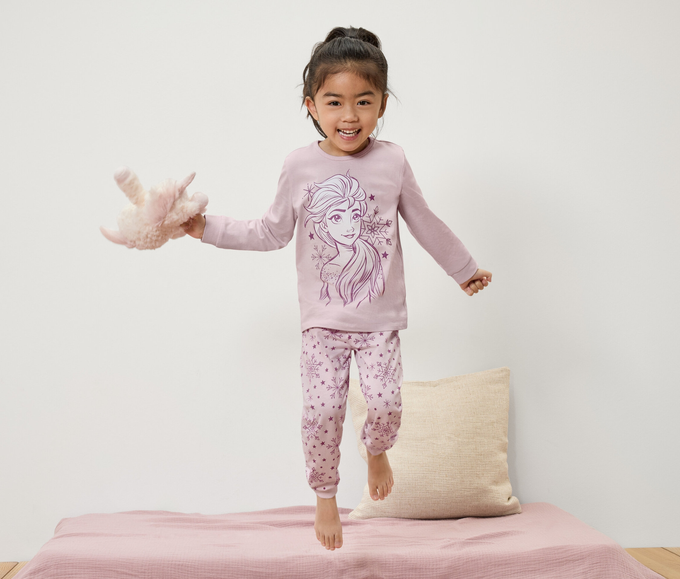 Fillette en pyjama Elsa sautant sur le lit tenant une peluche.