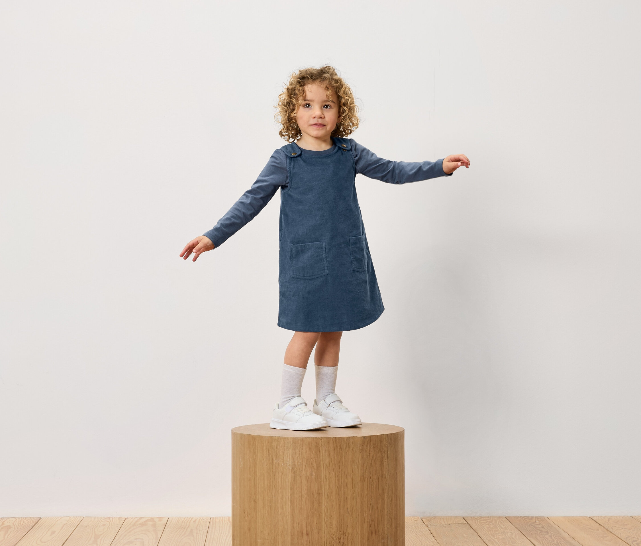 Une fille aux cheveux bouclés se tient sur un socle en bois. Elle porte une robe bleue et des baskets blanches.