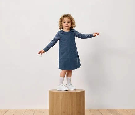 Une fille aux cheveux bouclés se tient sur un socle en bois. Elle porte une robe bleue et des baskets blanches.