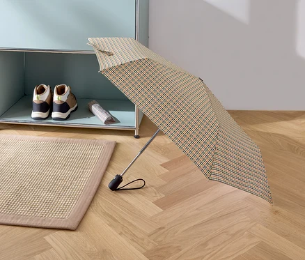 Un parapluie pliant ouvert à carreaux est posé sur un parquet. À l'arrière-plan, on voit un tapis beige, des chaussures et un sac sur une étagère.
