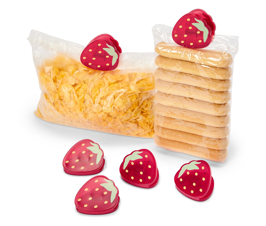 Un sac de flocons de maïs et un paquet de biscuits sont fermés par des clips en forme de fraise. Cinq autres clips en forme de fraise sont disposés au premier plan.