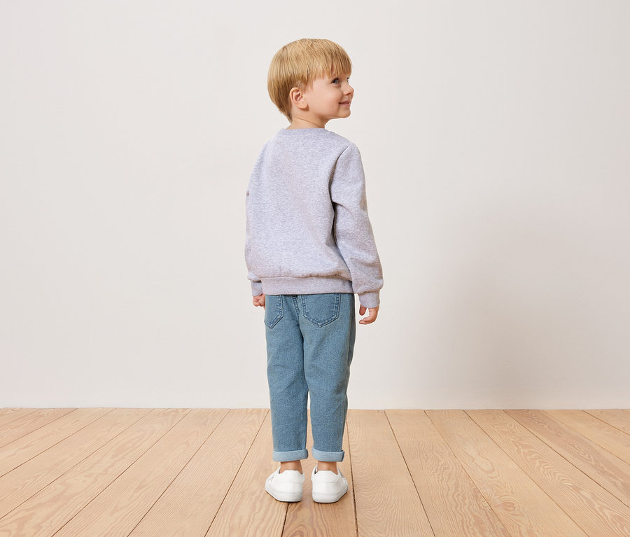 Petit garçon blond, vu de dos, portant un pull gris, un jean bleu et des chaussures blanches.