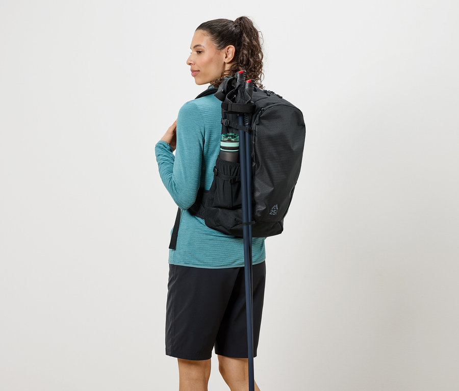 Femme vue de dos avec un sac à dos noir, une gourde et des bâtons de randonnée, vêtue d'un haut turquoise et d'un short noir.