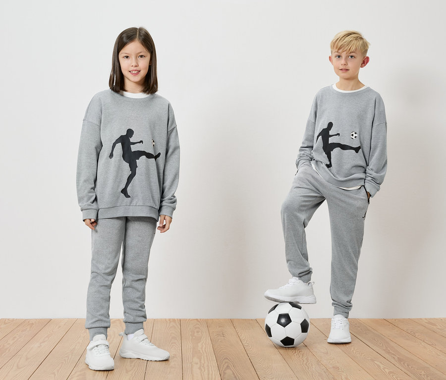 Deux enfants portent des survêtements gris avec des motifs de football. Un garçon a un pied sur un ballon de football.
