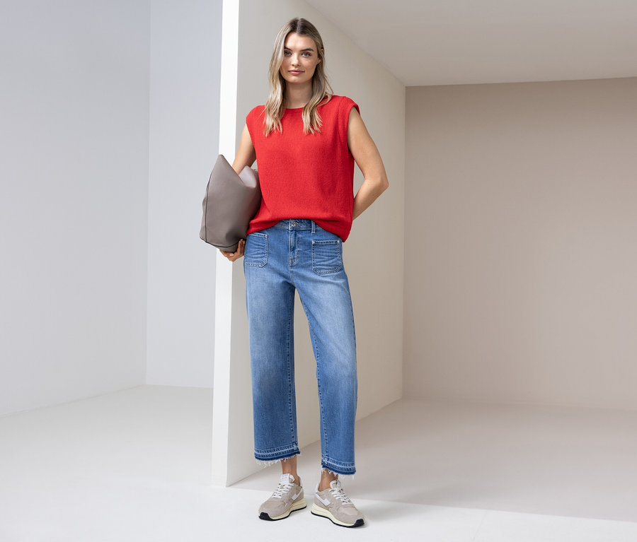 Une femme portant un haut rouge, un jean et des baskets se tient avec un sac gris dans une pièce lumineuse.