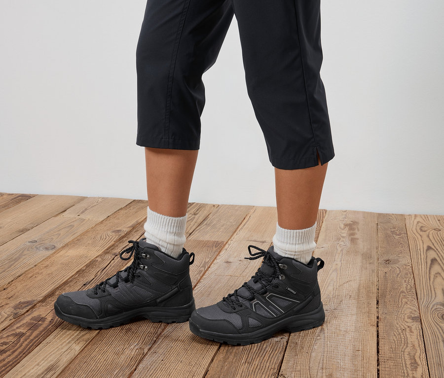 Une personne porte des chaussures de randonnée noires et un pantalon court sur un plancher en bois.