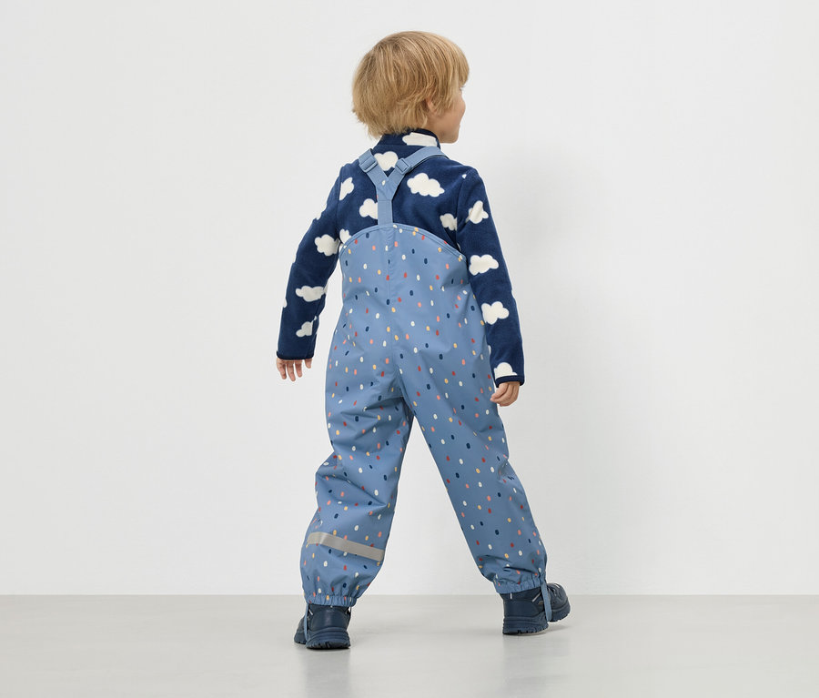 Enfant vu de dos, portant un pantalon de pluie bleu à pois colorés et un pull en polaire avec des nuages.