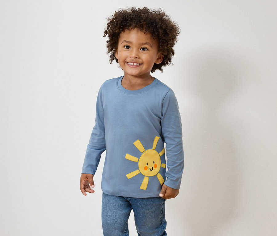 Un garçon souriant aux cheveux bouclés porte un t-shirt bleu à manches longues avec un appliqué de soleil jaune et un jean.