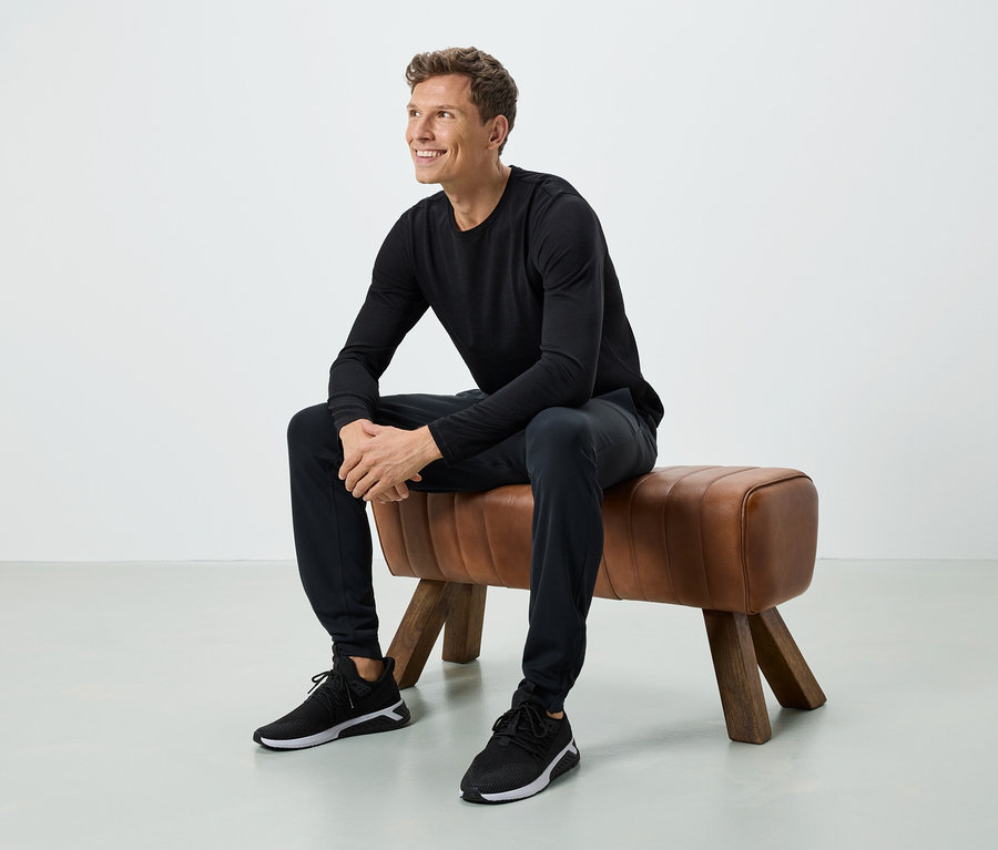 Un homme est assis sur un banc marron. Il porte un T-shirt noir à manches longues, un pantalon noir et des baskets noires.