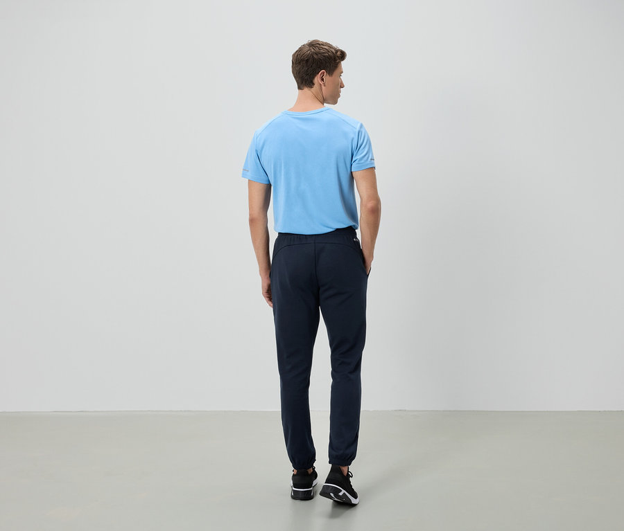 Un homme se tient dos à la caméra, portant un t-shirt bleu clair et un pantalon bleu foncé.