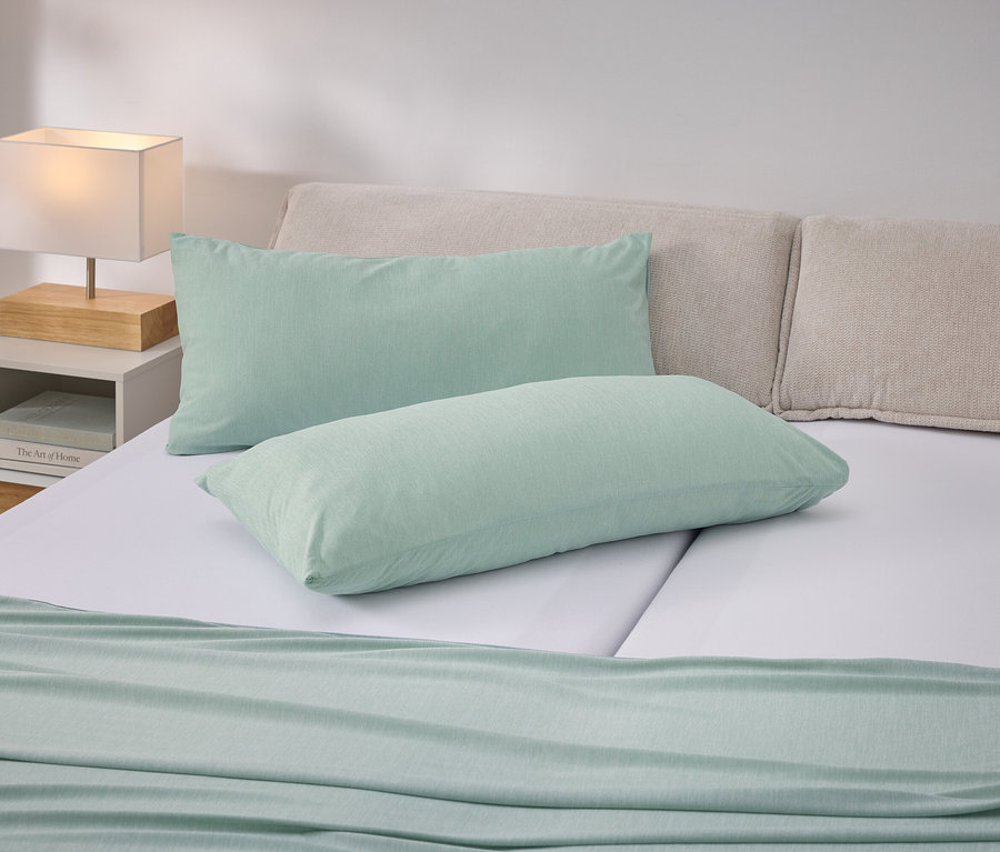 Lit avec coussins vert clair et couvre-lit sur des draps blancs. Table de chevet avec lampe à gauche.