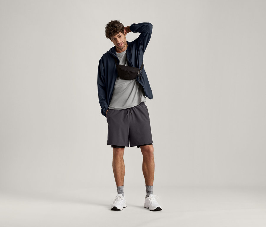 Un homme pose avec la main sur la tête, portant un t-shirt de sport gris clair chiné, une veste foncée, un short et un sac banane.