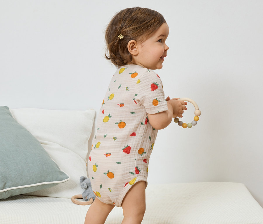 Une vue latérale d'un bébé avec une barrette brun clair, portant un body à manches courtes brun clair avec un imprimé de fruits et tenant un anneau de dentition en bois.