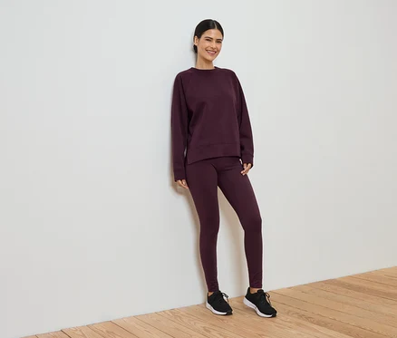 Une femme souriante en survêtement bordeaux composé d'un legging et d'un pull est appuyée contre un mur blanc.
