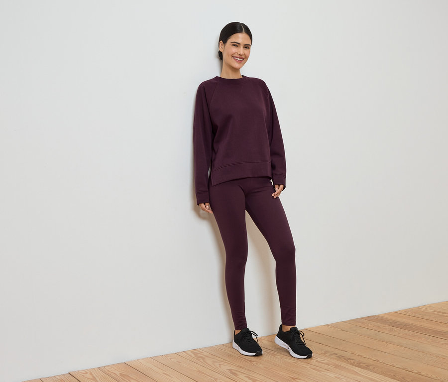 Une femme souriante en survêtement bordeaux composé d'un legging et d'un pull est appuyée contre un mur blanc.