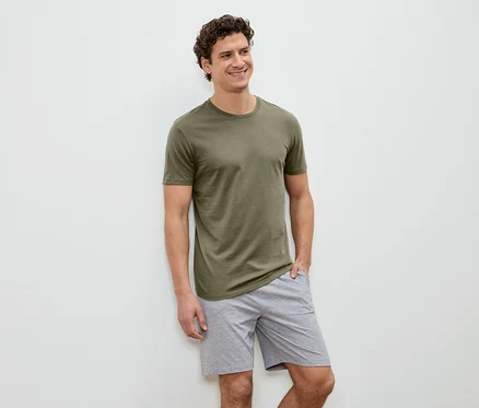 Un homme souriant aux cheveux bouclés, vêtu d'un t-shirt vert olive et d'un short gris, se tient devant un mur blanc.