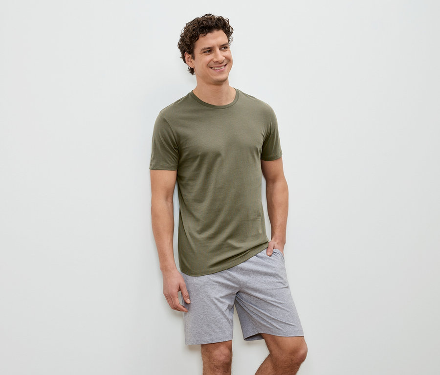 Un homme souriant aux cheveux bouclés, vêtu d'un t-shirt vert olive et d'un short gris, se tient devant un mur blanc.