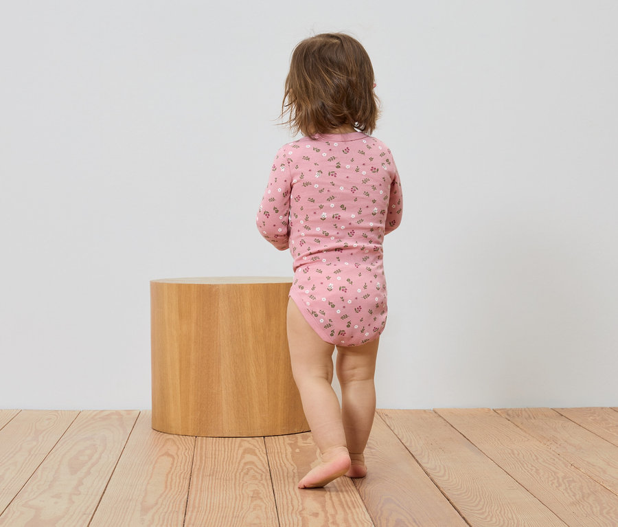 Un enfant en body rose à fleurs se tient dos à un tabouret cylindrique marron clair.