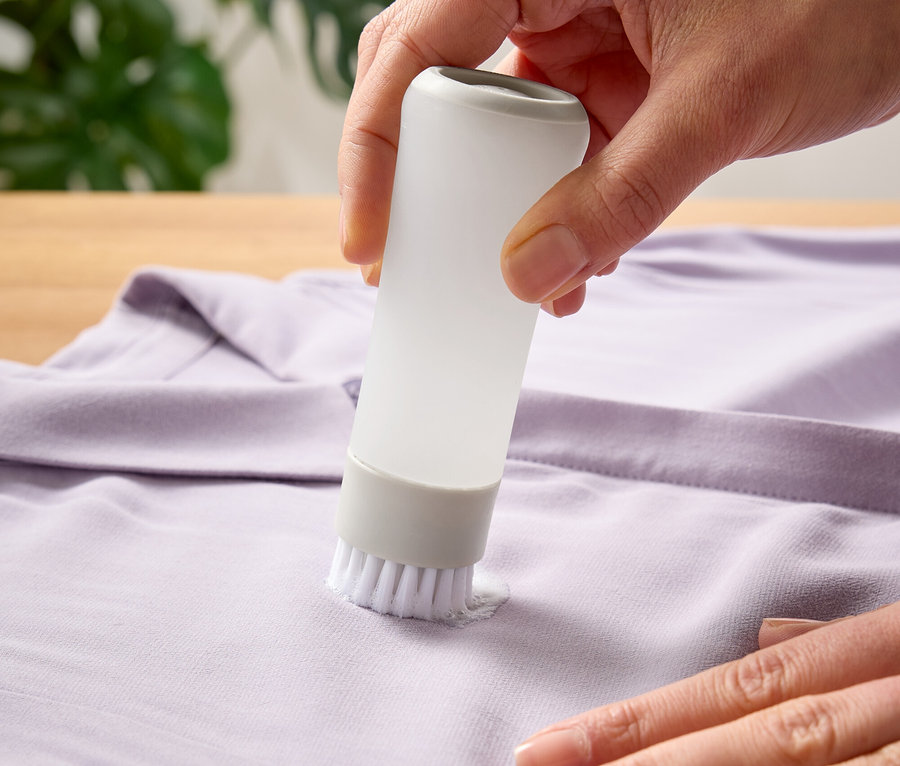 Une main utilise une brosse avec réservoir pour traiter une tache sur un t-shirt violet.