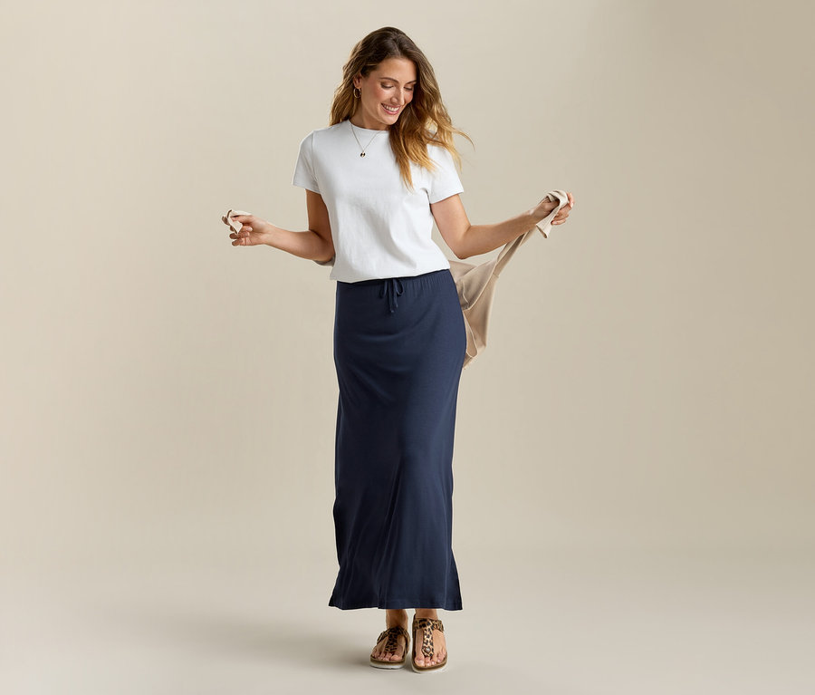 Une femme tient un cardigan beige, porte un t-shirt blanc et une jupe longue en jersey bleu marine.