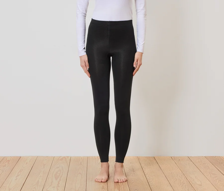 Une personne pieds nus portant un haut blanc à manches longues et un legging noir se tient devant un mur et un plancher en bois.