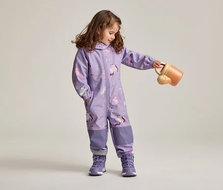 Une fillette aux cheveux bruns et bouclés, vêtue d'une combinaison violette à motifs de licornes et de chaussures violettes, tient un arrosoir orange.