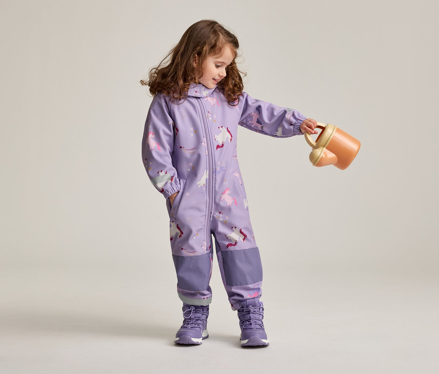 Une fillette aux cheveux bruns et bouclés, vêtue d'une combinaison violette à motifs de licornes et de chaussures violettes, tient un arrosoir orange.